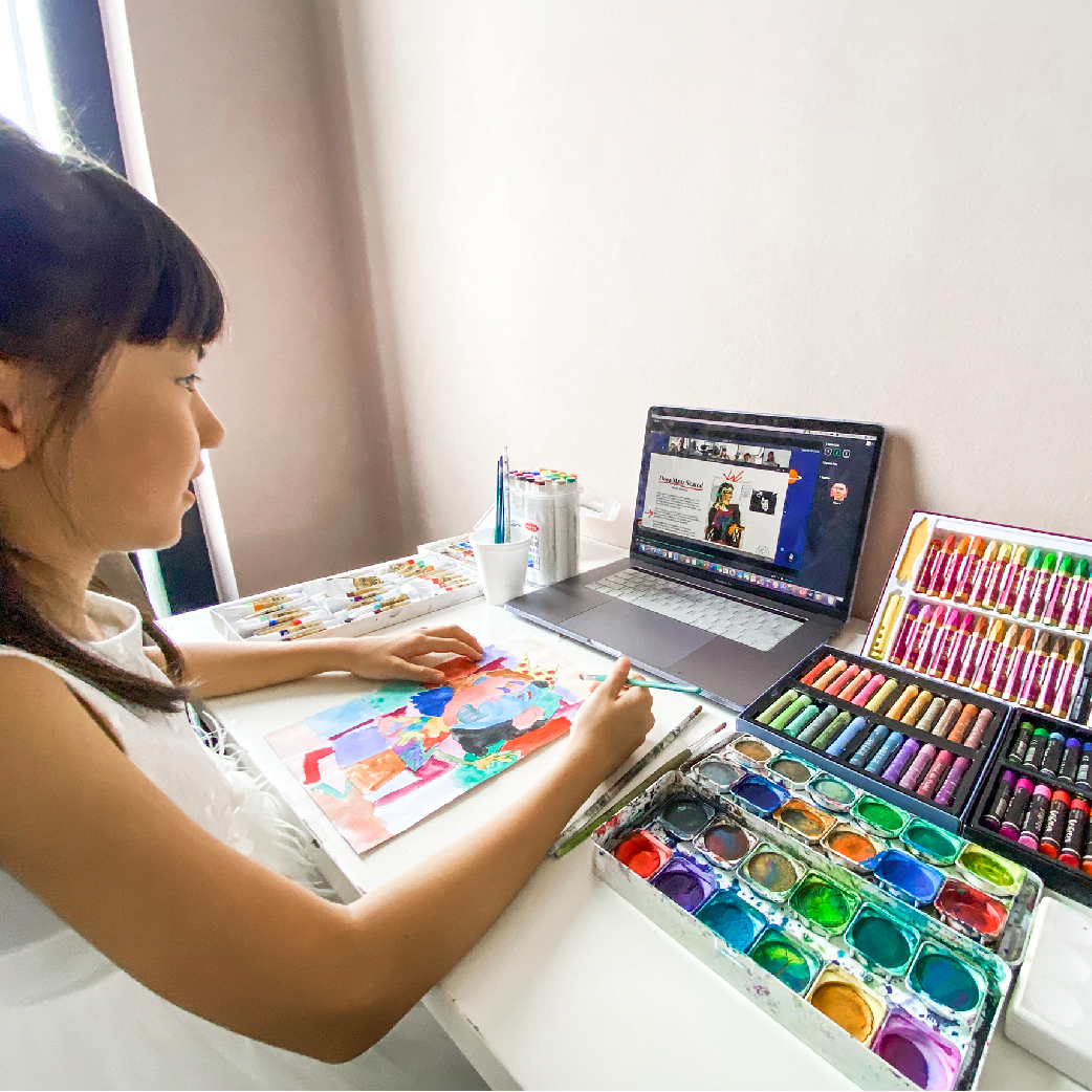 Girl painting at a home desk beside a laptop streaming a live online art lesson, watercolours and oil pastels within reach