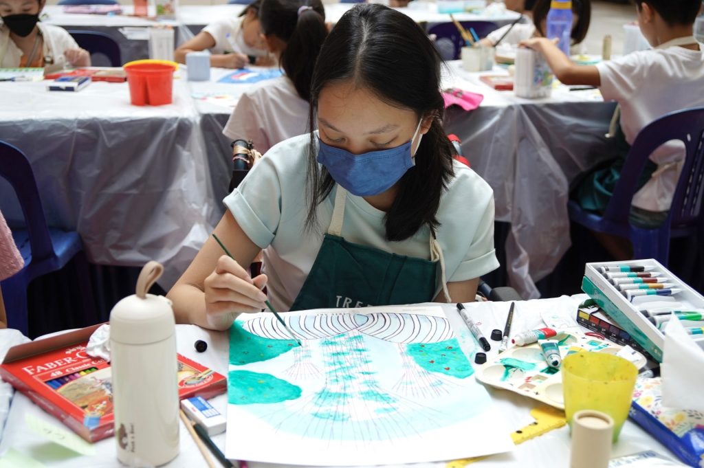 Young artist in a TreeArt apron painting a watercolour piece at a group table during the Kine x CultuRediscover art competition