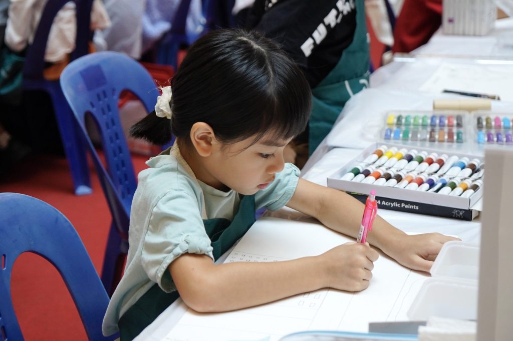 Young girl concentrating on a drawing exercise at a classroom table, with a set of acrylic paints beside her worksheet