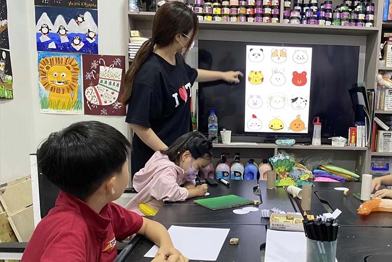 Teacher demonstrating digital character design on a screen while children work on crafts at the table