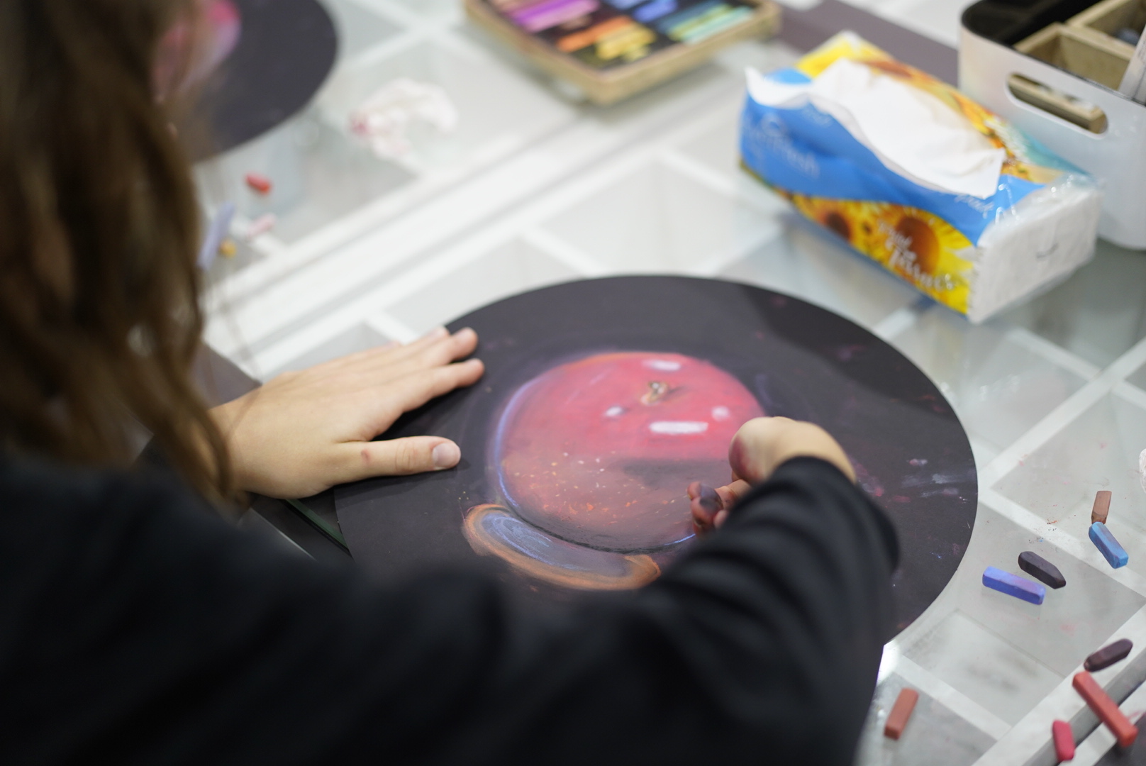 Close-up of a child's hands blending a red soft-pastel apple on black paper