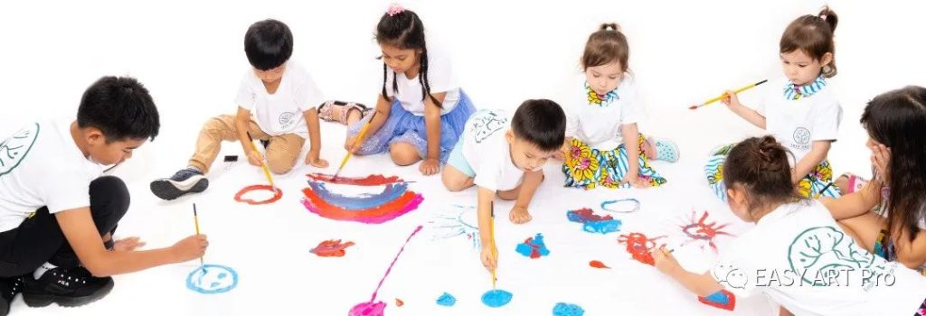 A group of children in Tree Art t-shirts sitting together on the floor, freely painting on a large shared canvas with colorful paints
