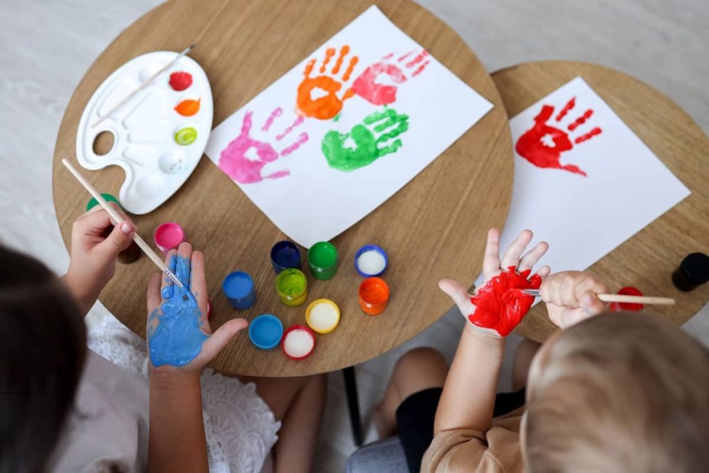 Two children making colourful handprint paintings, with paint pots and a palette laid out on the table