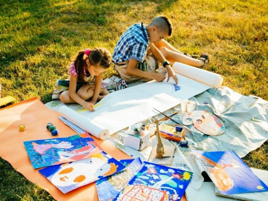 Two children painting together on a large roll of paper outdoors in a sunny, grassy field