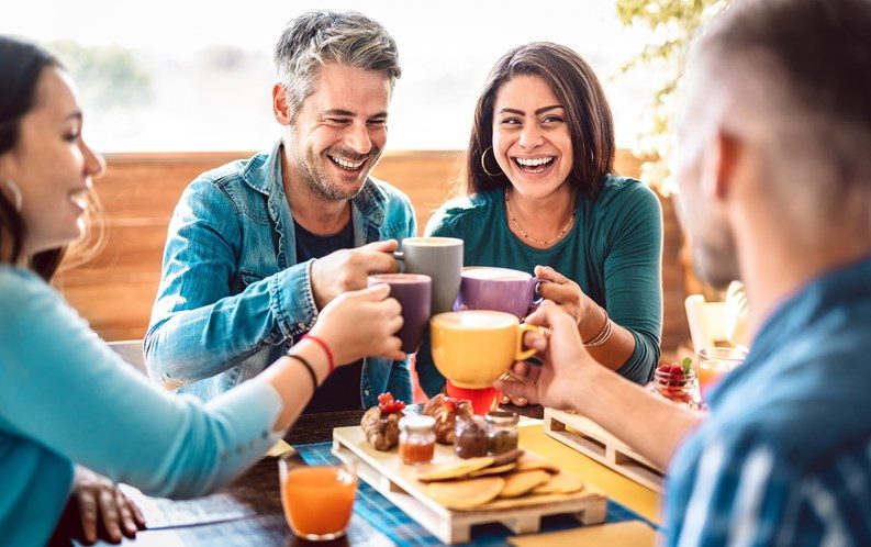 A group of friends laughing together and toasting mugs at a café table
