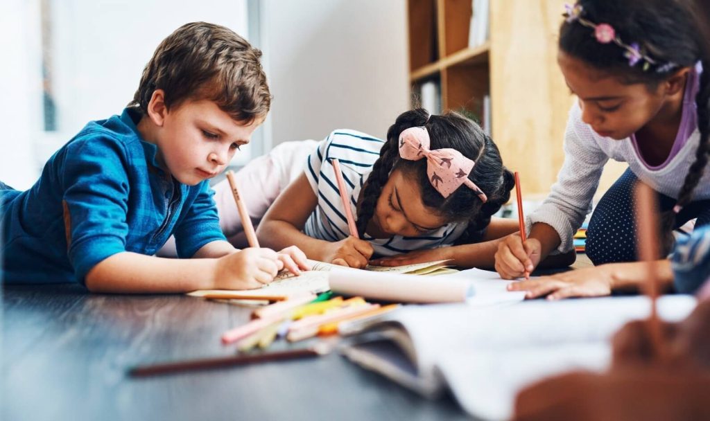 Three children lying on the floor together, focused on drawing with colored pencils
