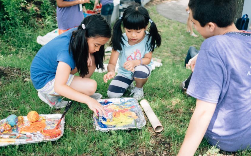 Children kneeling on grass outdoors, painting together on foil trays