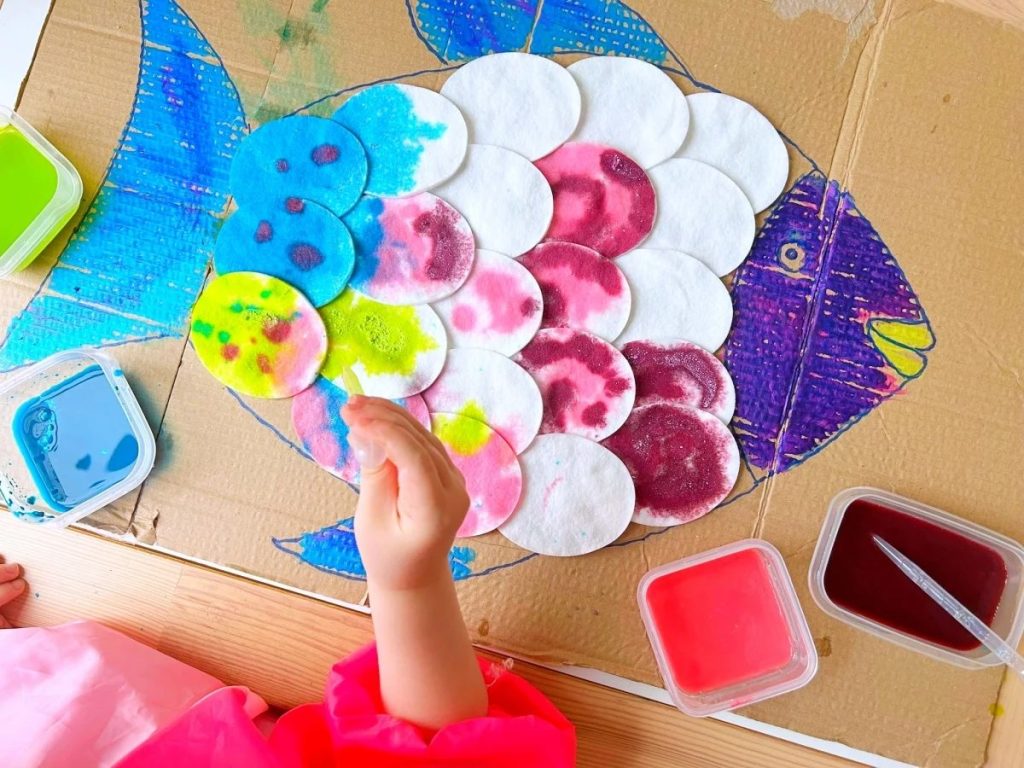 Child's hand carefully placing painted cotton pad scales onto a cardboard fish craft, practising fine motor control