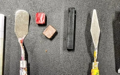 Close-up of assorted art tools and materials, including palette knives, an oil pastel and a charcoal stick, laid out for experimentation