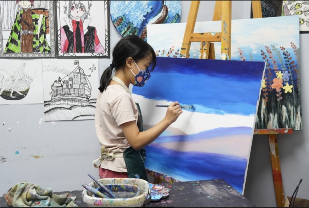 Student at Tree Art painting a blue seascape on canvas at an easel, surrounded by other students' artwork in the studio