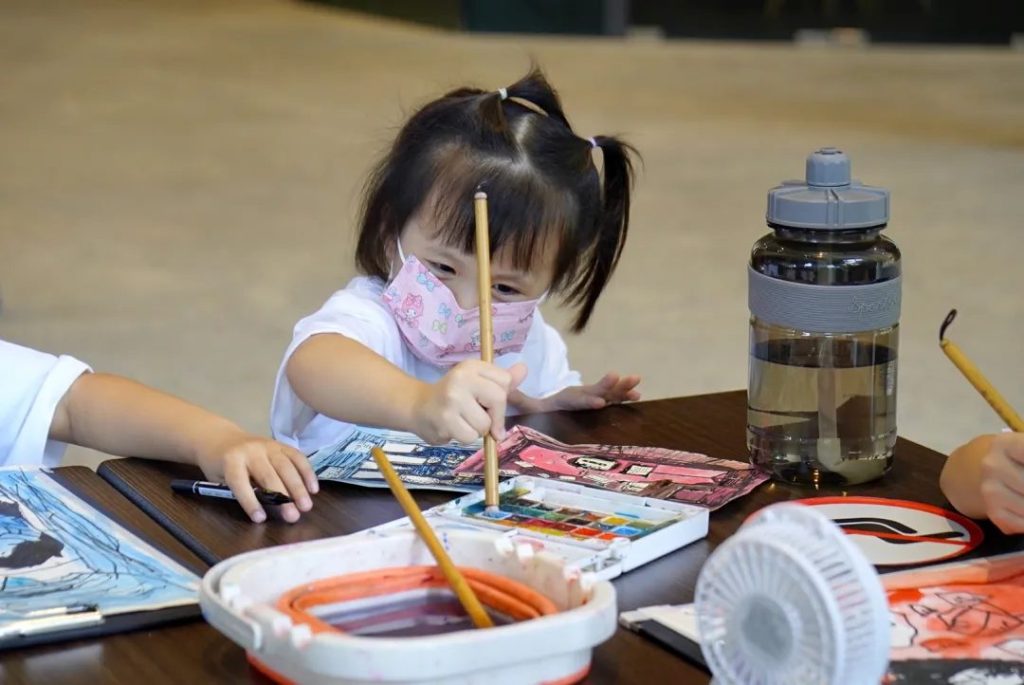 Young girl in pigtails and a face mask painting with watercolours at a table