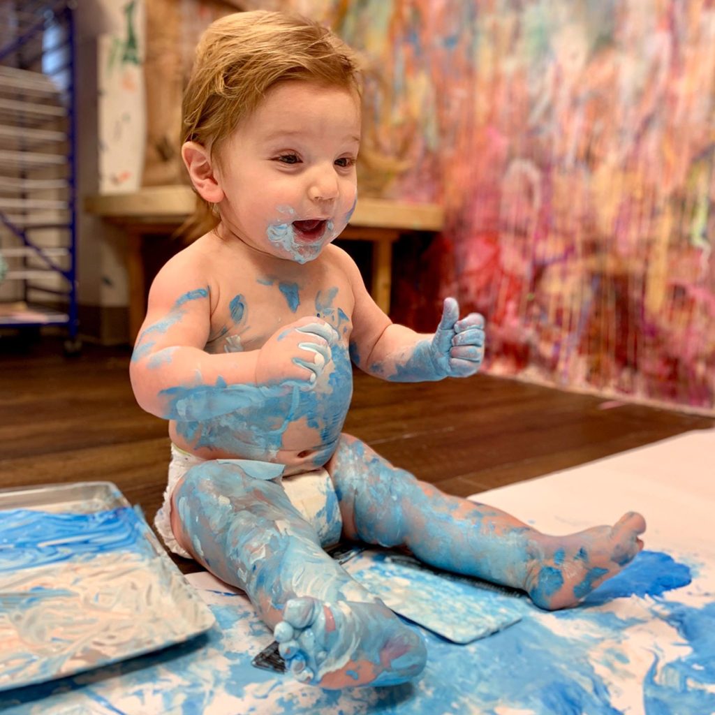 Toddler joyfully covered in blue paint during a hands-on art class, exploring creativity through sensory play