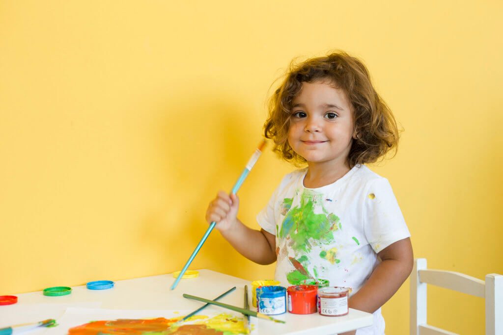Smiling child holding a paintbrush with colourful paint on her white shirt