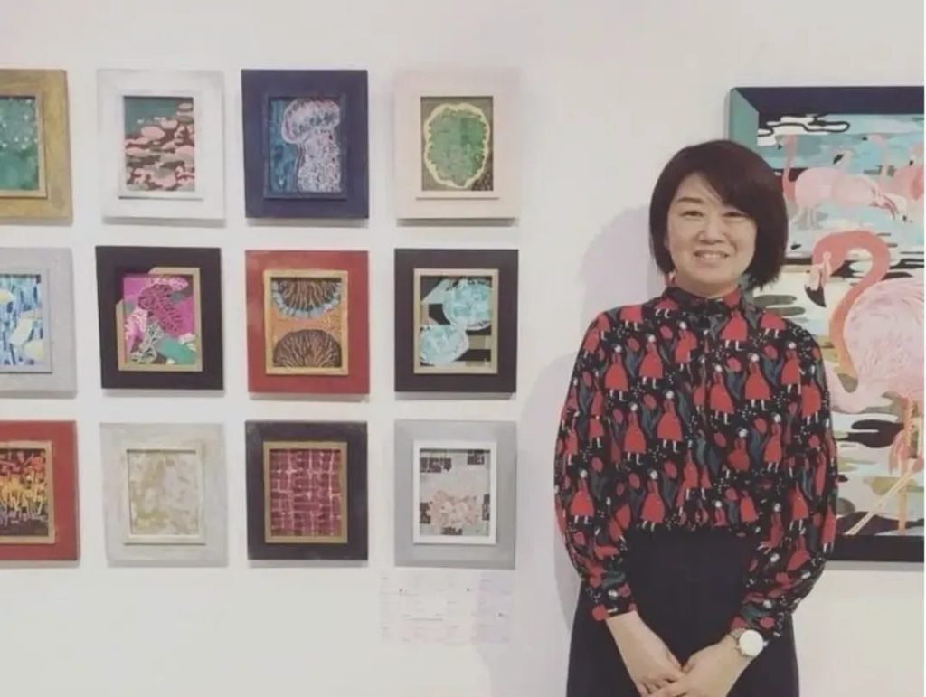 Yuko Kurihara standing beside a gallery wall of her framed Nihonga paintings, with a large flamingo-themed artwork beside her