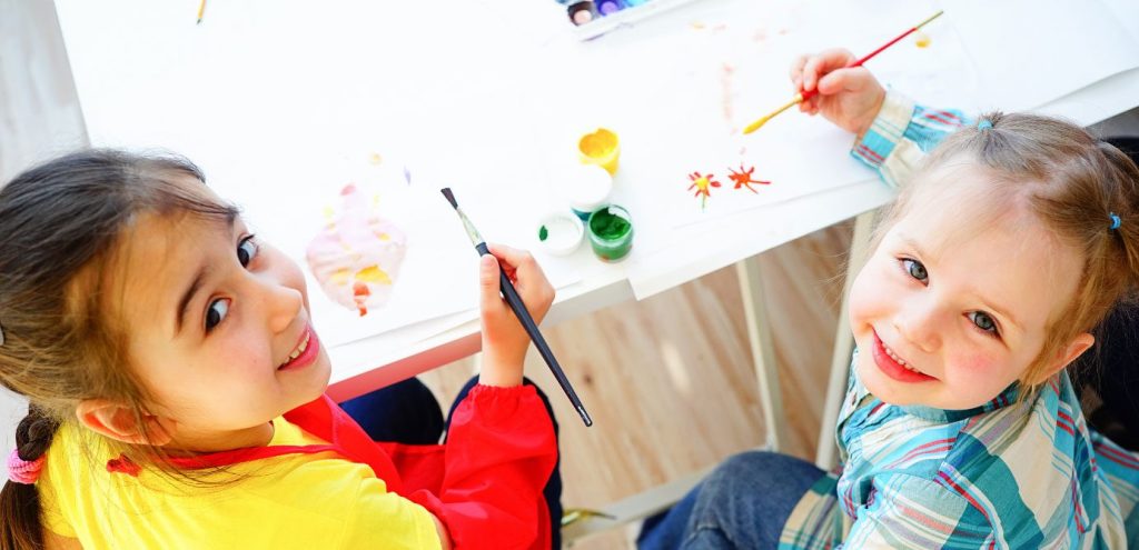 Two young girls smiling and painting together at a shared table