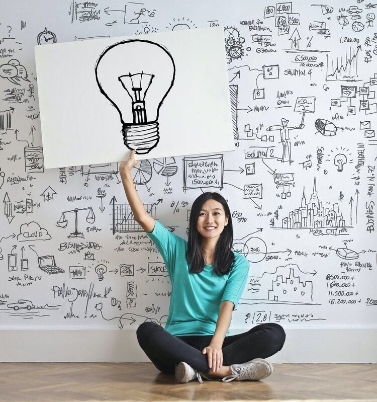 A person holding up a hand-drawn lightbulb sketch in front of a wall covered with idea diagrams and charts