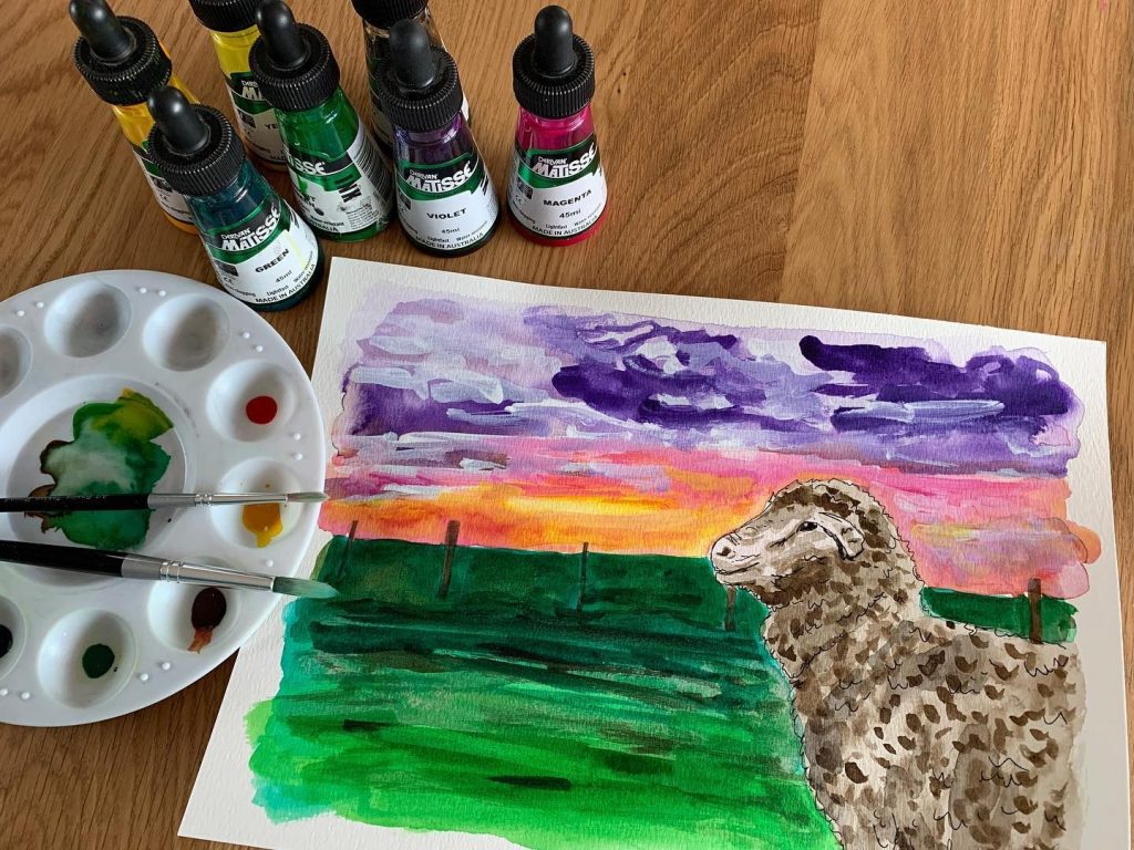 Watercolour painting of a sheep in a field under a sunset sky, surrounded by paint bottles, a palette, and brushes