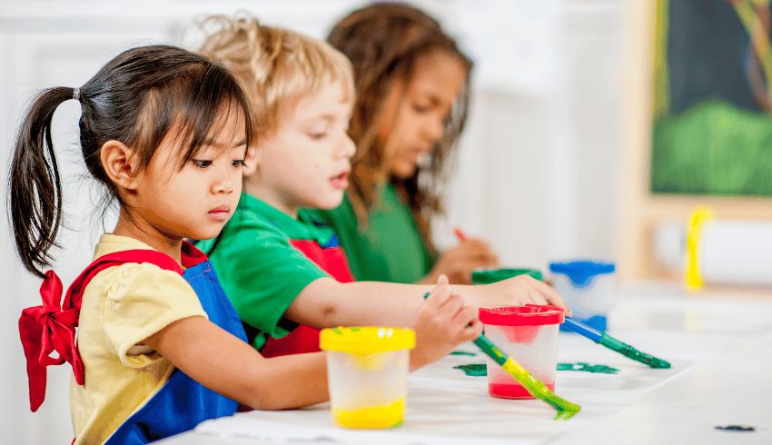 Three young children in aprons painting together at a table