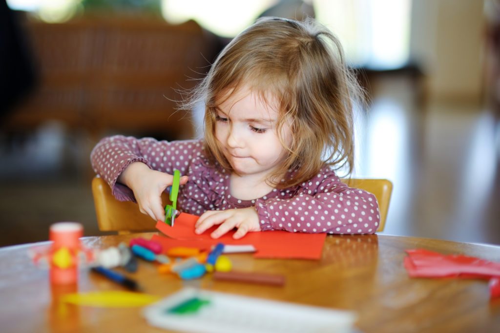 A young girl carefully cutting red paper with scissors at a table covered in craft supplies