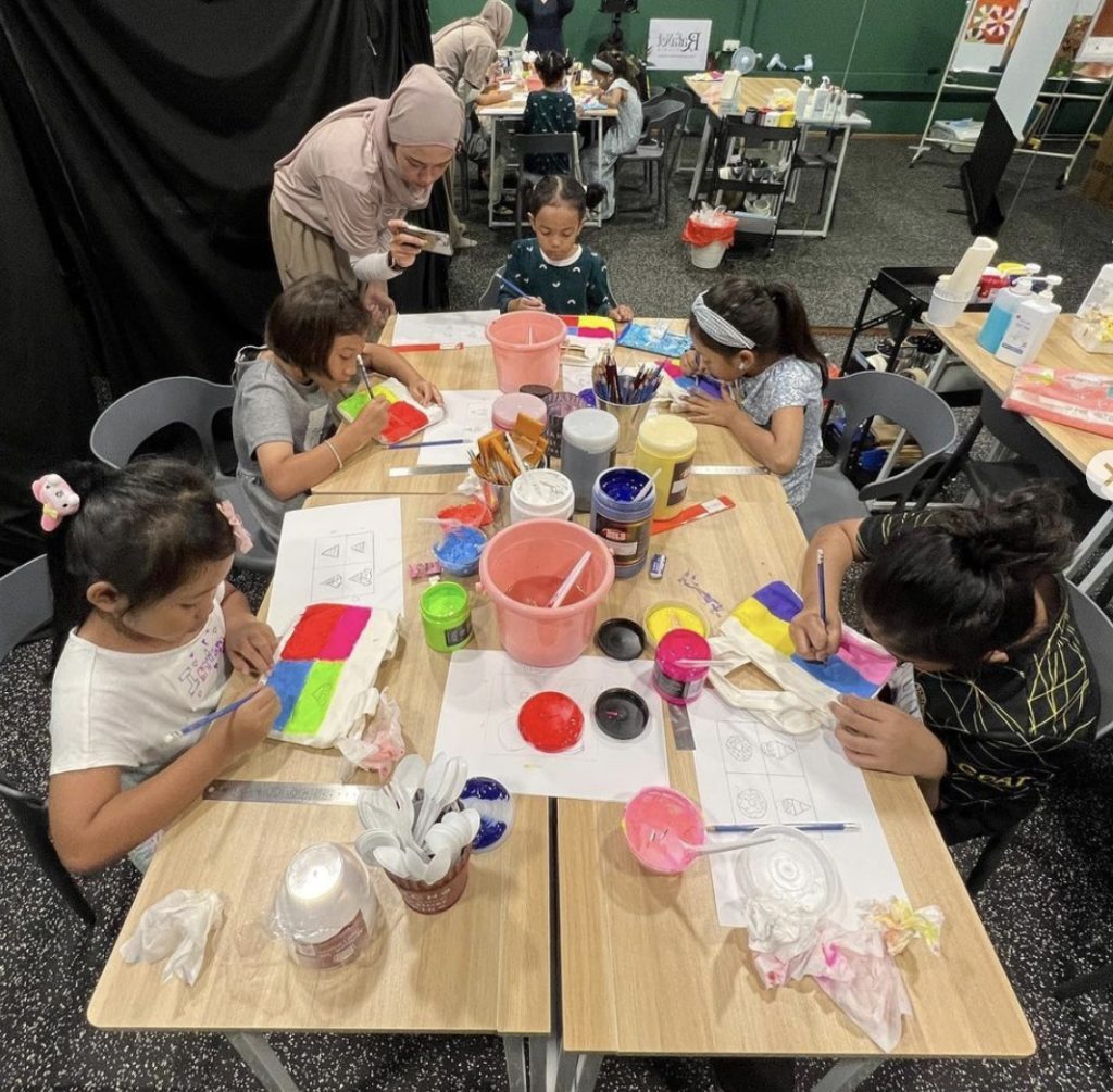Art instructor guiding children as they paint tote bags at a classroom table filled with paint and brushes