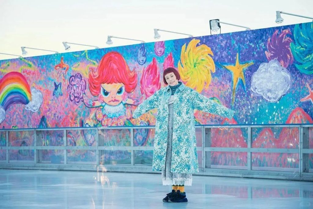 Bibi Lei posing in front of a large outdoor mural of her signature red-haired girl character at an exhibition