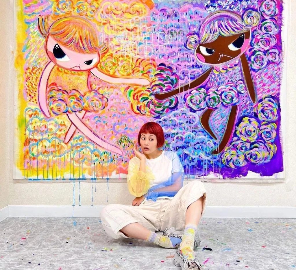 Bibi Lei sitting in her studio in front of a large mural of two of her signature girl characters reaching toward each other