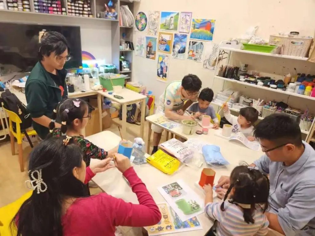Teachers and parents guiding children as they paint clay cups together at a table in a TreeArt studio surrounded by diverse student artwork