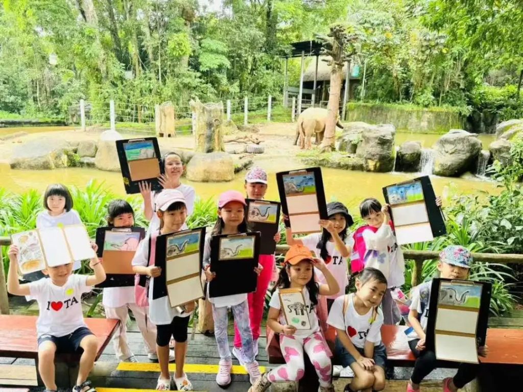 Children sketching elephants from observation on an outdoor zoo excursion, each holding up their own drawing on a clipboard