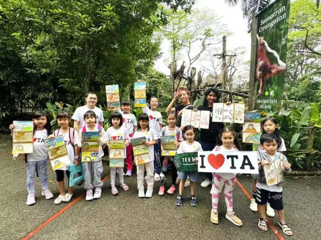 TreeArt students and teachers holding their zoo-themed artwork beside a Sumatran Orangutan exhibit sign and an "I love TA" banner at Singapore Zoo