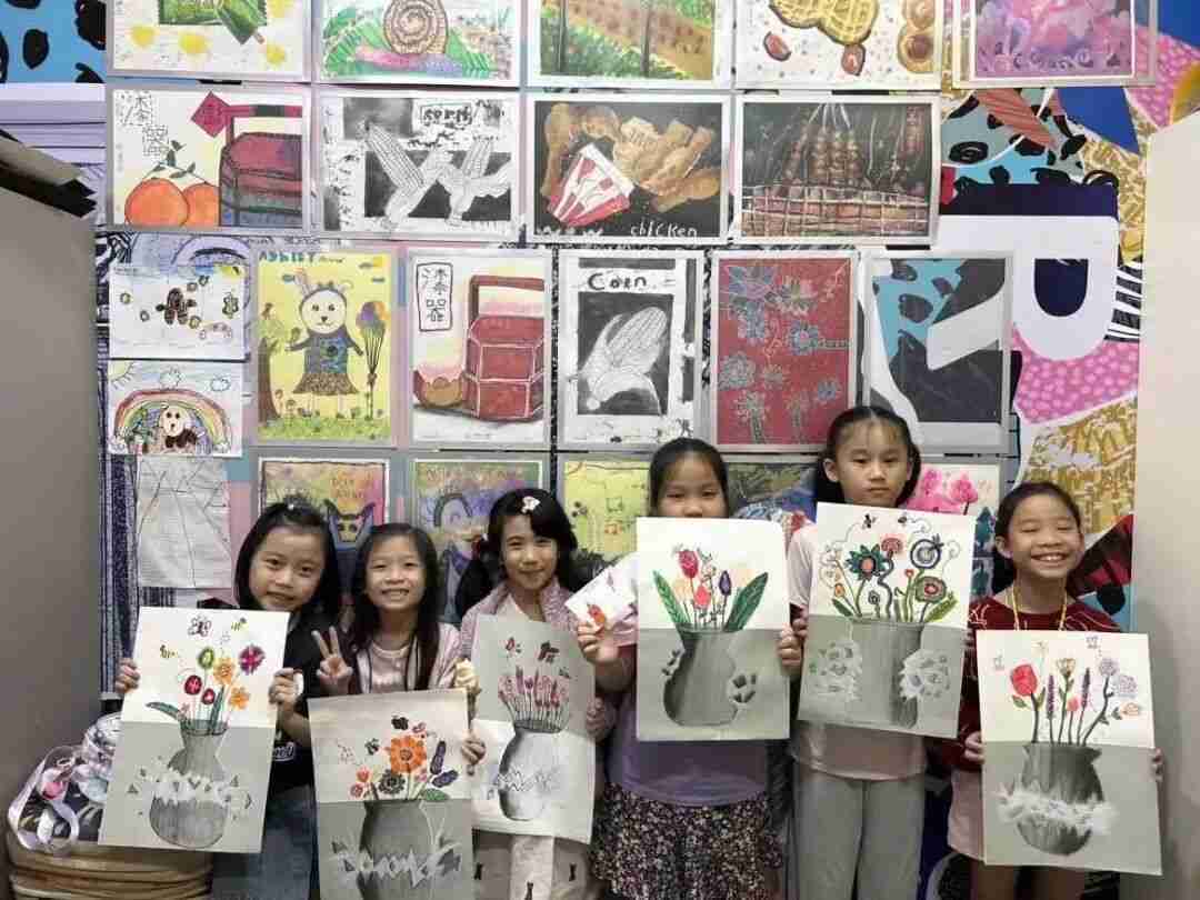 Children holding up their flower-vase paintings in front of a wall of classroom artwork