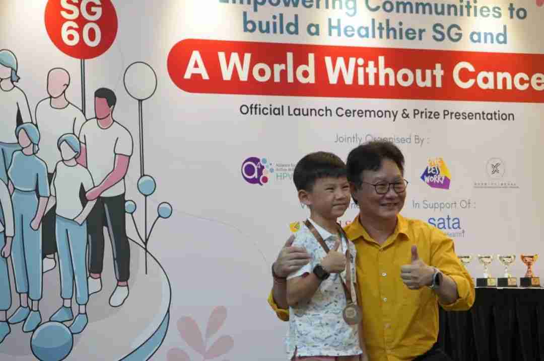 Guest speaker Mr Koh Juay Meng poses with a young medal-winning participant in front of the SG60 "World Without Cancer" ceremony backdrop