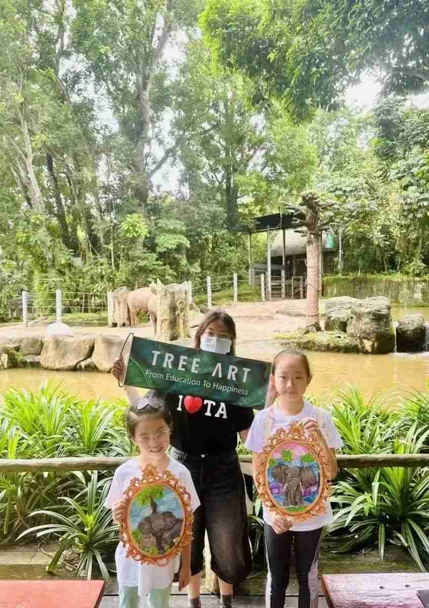 TreeArt students holding their framed elephant paintings at a zoo, with a TREE ART banner and elephants in the background