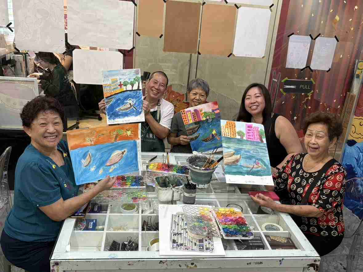 Adult and senior students around a shared studio worktable, laughing as they hold up the watercolour boat and harbour scenes they have just finished