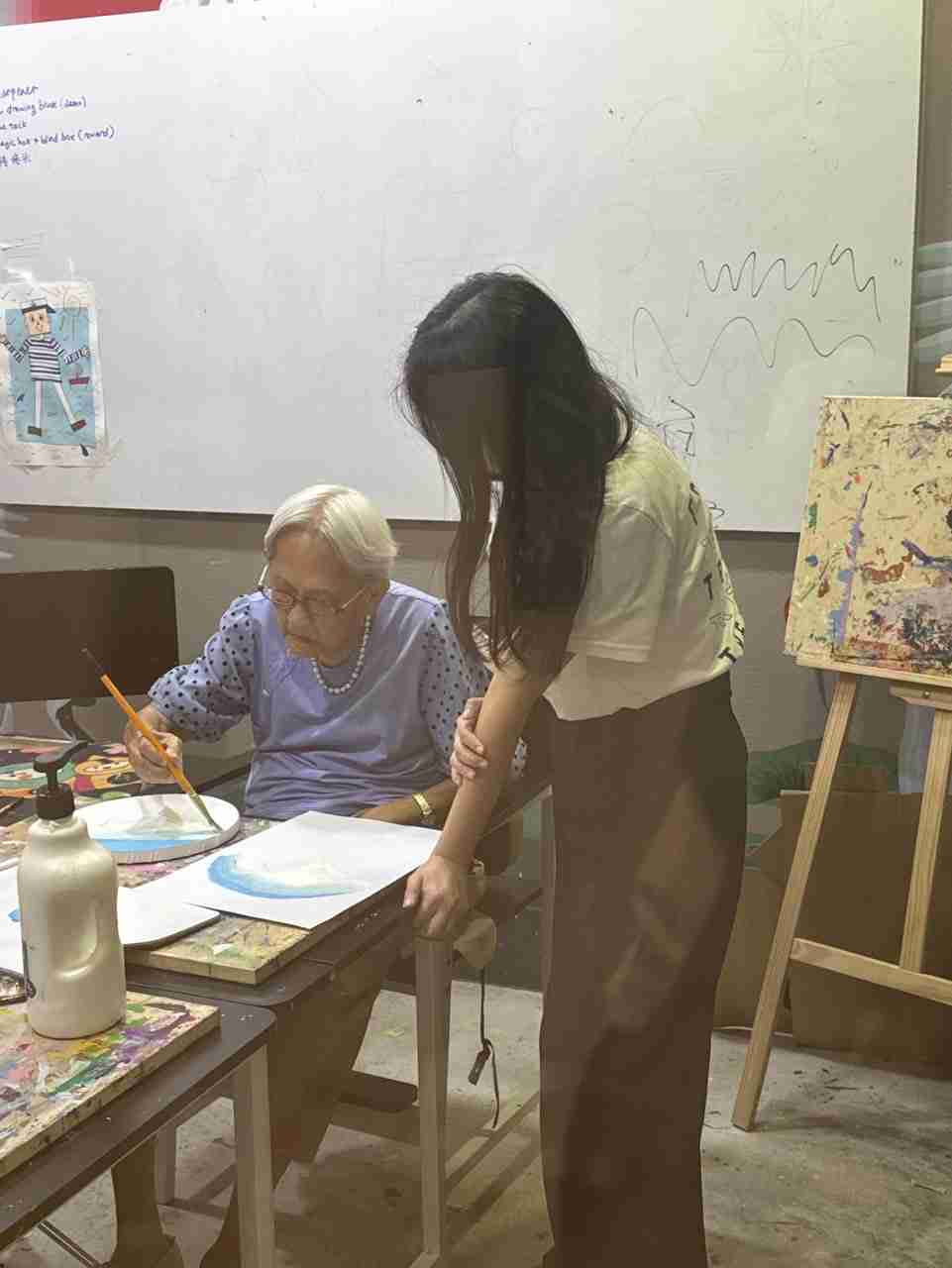 Teacher guiding a senior student through a painting at a studio table