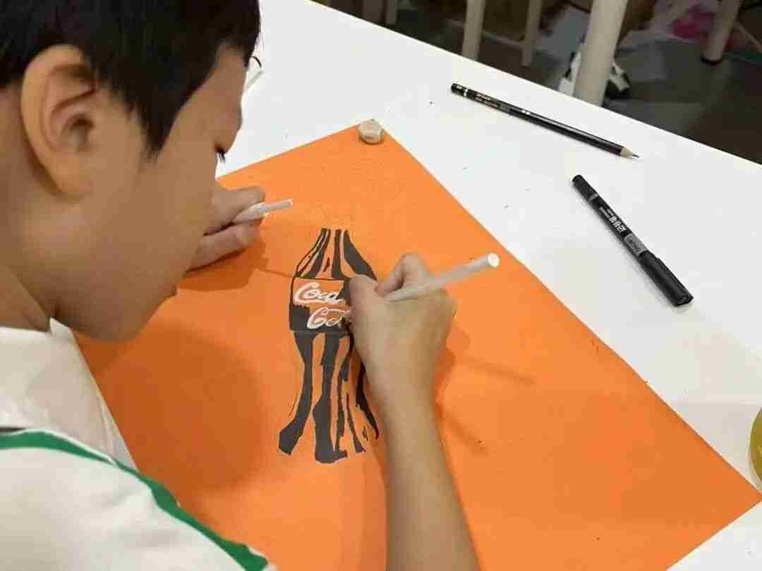A child carefully sketching a Coca-Cola bottle in pencil on orange paper