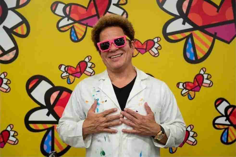 Romero Britto in pink glasses and a paint-stained coat in front of a yellow wall of his winged-heart motifs