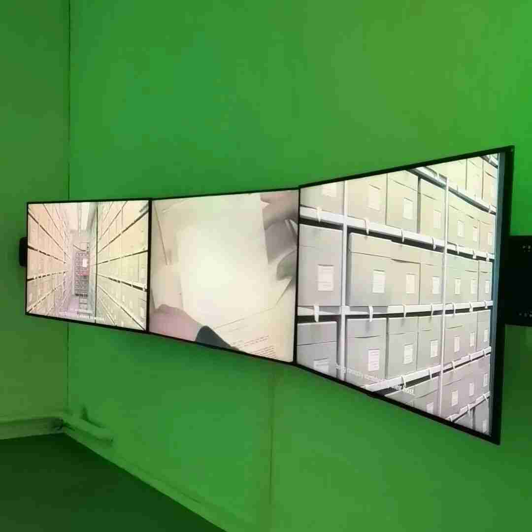 Three-panel video installation showing archival footage of storage boxes, mounted on a green gallery wall