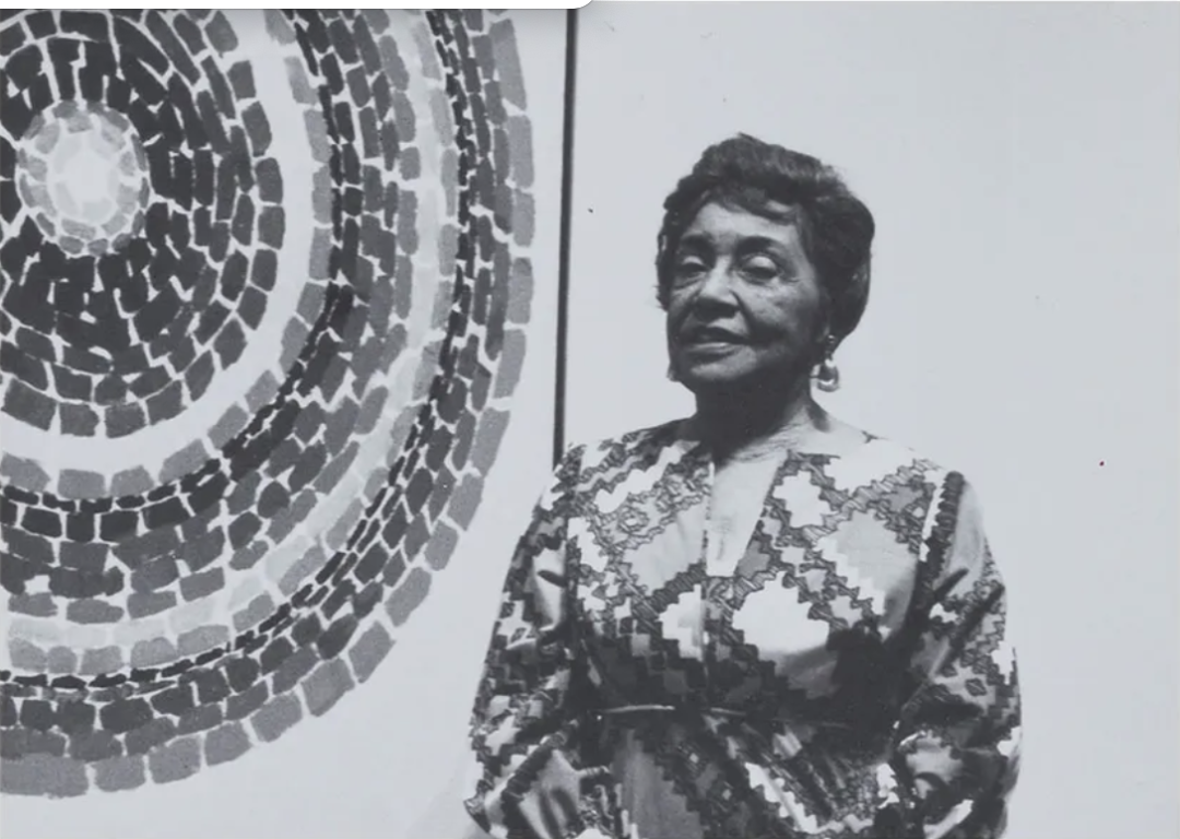 Black-and-white photograph of artist Alma Thomas standing beside one of her circular mosaic-patterned paintings