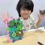 Child proudly holding up a handmade 3D craft featuring a garden scene and a decorative hand shape