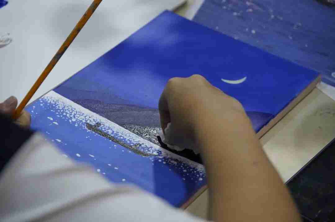Child's hand painting a deep blue night sky with a crescent moon and sea using a fine paintbrush