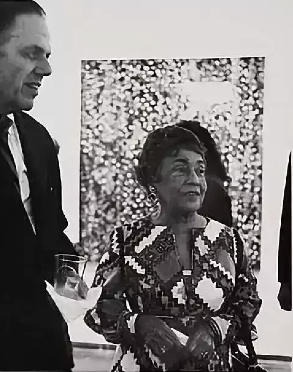 Black-and-white photograph of Alma Thomas at a gallery opening, speaking with a man in a suit, one of her dotted paintings displayed behind them