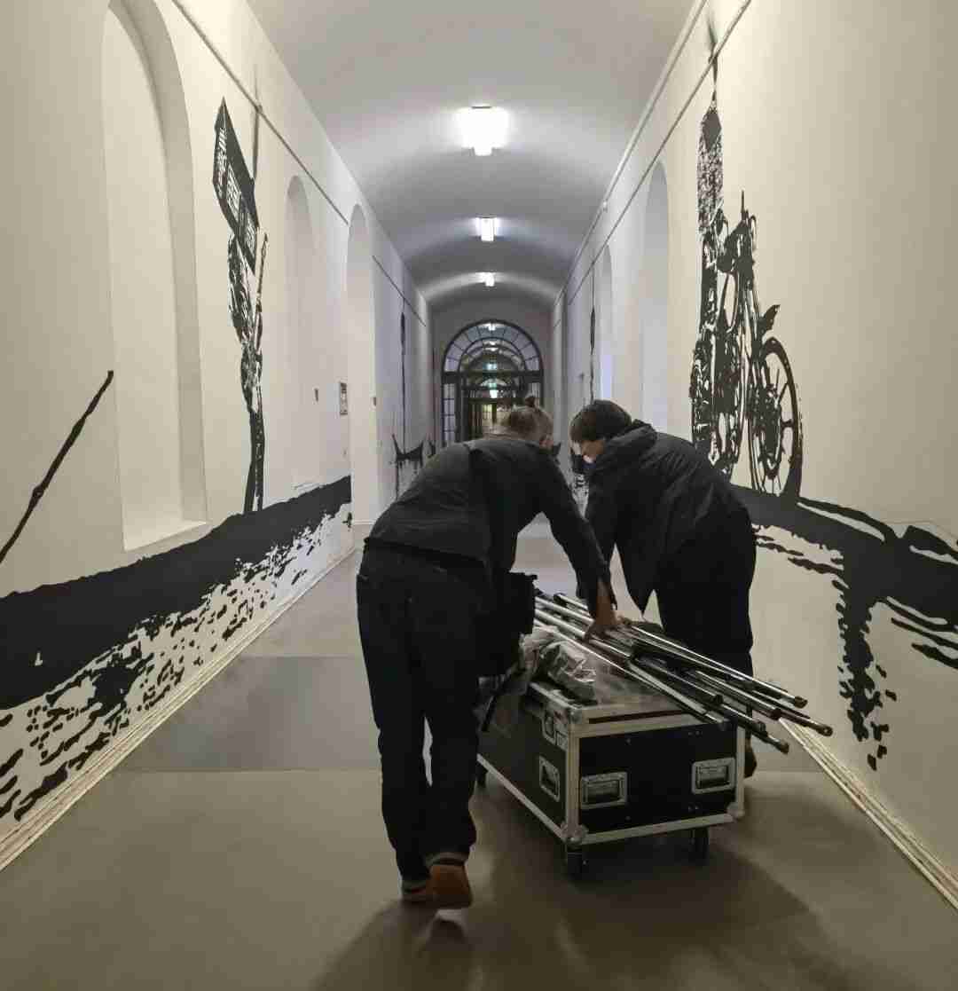 Installation crew moving equipment through a vaulted gallery corridor lined with large black-and-white mural drawings