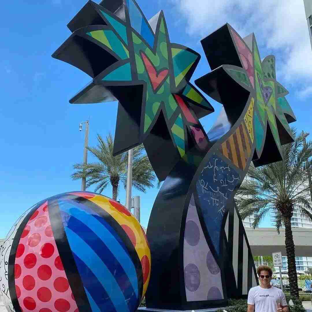 Romero Britto standing beneath his monumental outdoor sculpture among palm trees