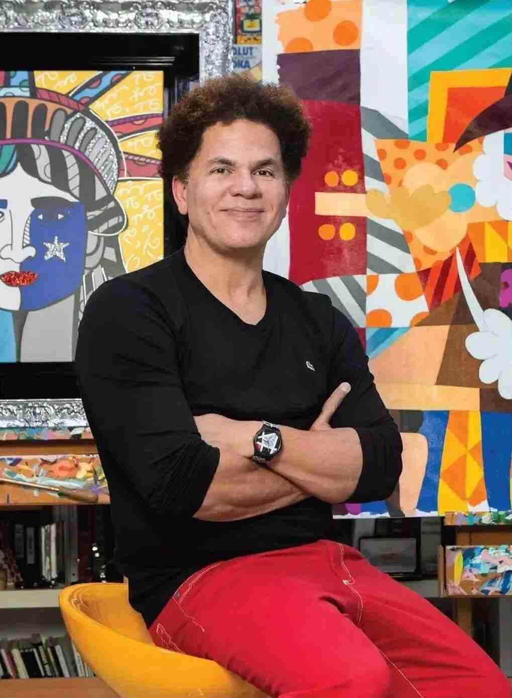 Romero Britto seated in his studio with arms crossed, surrounded by his artworks