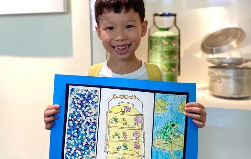 A smiling boy holds up his framed drawing of a tiered tiffin carrier in a museum