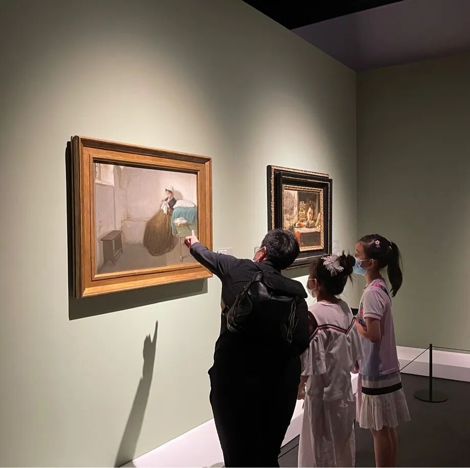 An adult points at a classical painting while two young girls look on in a museum