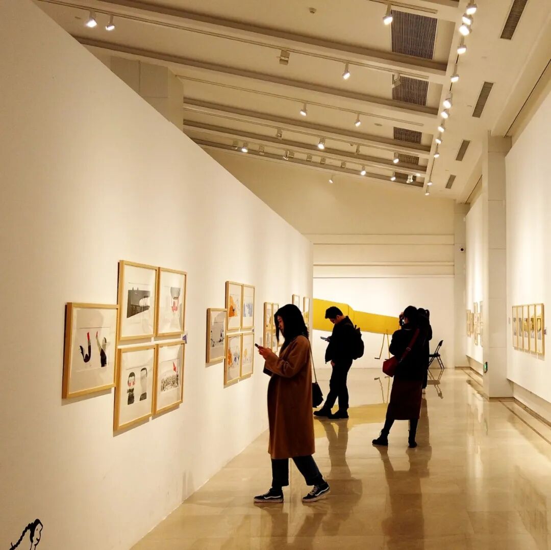 Visitors walk through a warmly lit gallery corridor hung with framed illustrations