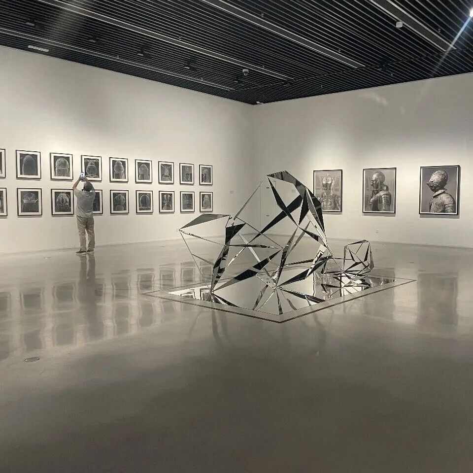 A mirrored geometric sculpture installation in a gallery lined with black-and-white photographs