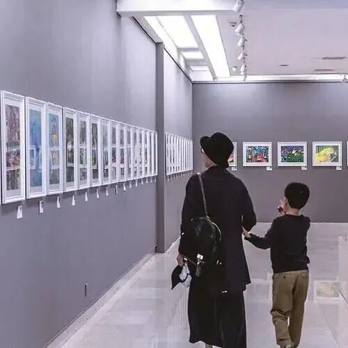 A parent and child walk hand in hand through a gallery lined with framed children's paintings