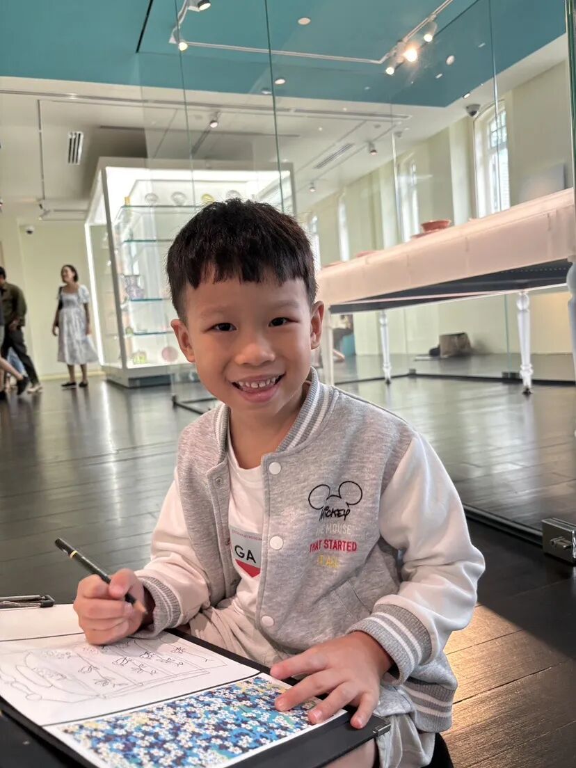 A smiling boy sketches on a clipboard while sitting on the floor of a museum gallery